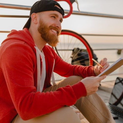 handsome-hipster-style-bearded-man-studying-online-freelancer-writing-making-notes-with-backpack-bicycle-morning-sunrise-by-sea-healthy-active-lifestyle-traveler-backpacker